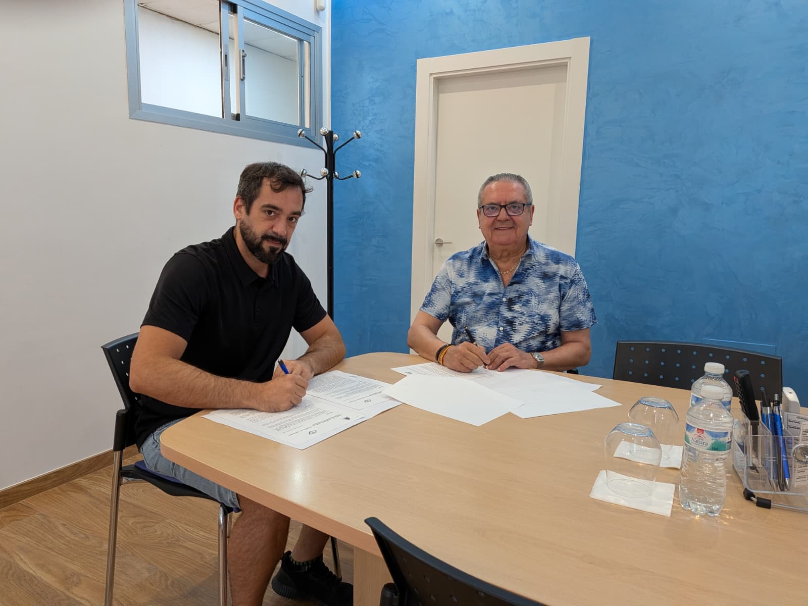 Dos hombres sentados a una mesa de madera firmando el convenio con Promanimport y la ACET. A la izquierda, un hombre joven con polo negro sostiene un bolígrafo azul; a la derecha, un hombre mayor con camisa azul estampada sonríe frente a una pared azul. Convenio proman Import